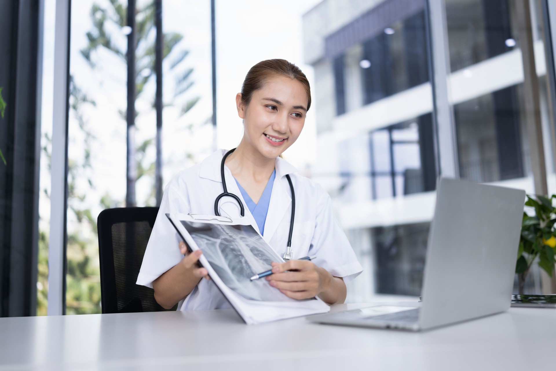 Happy smiling female asian doctor working with patient x-ray film and laptop computer sitting at desk in hospital.